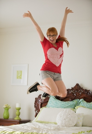Excited redhead needs somewhere around just jumping on bed so she unclothes before window